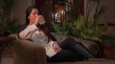 Kendine güvenen, resmi kıyafetleri olan genç bir kadın kanepede oturur ve şık bir salonda şık bir şekilde viskisini yudumlar. İç mekan bitkileri, hafif ışıklandırma ve rahat profesyonel atmosfer.