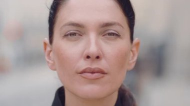 Tarafsız ifadesi olan, saçları arkadan bağlı, doğal makyajlı, doğrudan kameraya bakan, gün ışığı ve sığ alan derinliğindeki yumuşak kentsel geçmişe sahip genç bir kadının yakın görüntüsü.