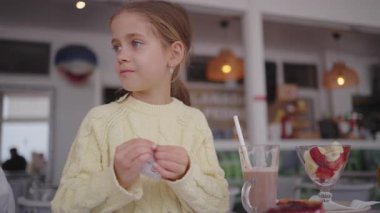 Kafe masasında şeker kesesini açmaya çalışan sarı kazaklı tatlı kız. Bir bardak çikolatalı içecek ve önünde meyve ve krema olan tatlı kasesi. Günlük kafeterya atmosferi