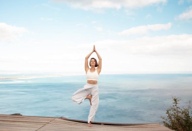 Spor giyim bölümünde formda bir kadın, ağaç poz yogasını deniz kenarında ahşap platformda yapıyor. Bayan dengeyi koruyor ve zarafetle geriniyor doğa ve sükunetle çevrili.