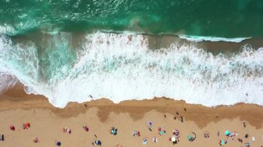 Güneşli bir günde, turistlerin kumlu deniz kıyısında güneşli bir sahil şeridinde güneşlenen ve dalgaların kıyıya vurduğu hava aracı görüntüsü.