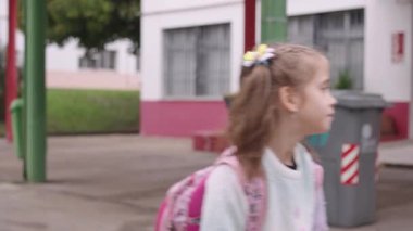 Sırt çantalı küçük kız gündüz vakti sokakta yürüyor, neşeyle gülümsüyor. Parlak ve canlı bir ortamda yürürken mutlu görünüyor.