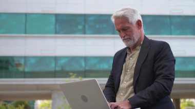 Kendine güvenen kıdemli işadamı ofis binasının yanında dizüstü bilgisayar kullanıyor. Gündüz vakti modern şehir ortamında çalışıyor.
