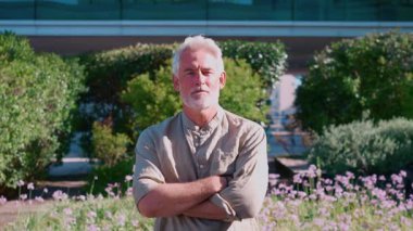 Beyaz saçlı ve sakallı kendinden emin işadamının modern ofis binası ve yeşillik yakınlarında kolları çaprazlanmış şekilde görüntüsünü yakınlaştır.