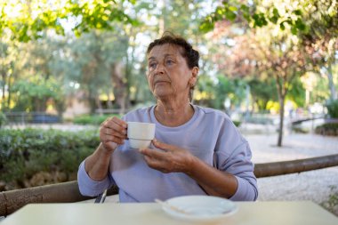 Kaldırımdaki kafede elinde kahve fincanı tutan düşünceli yaşlı kadın yeşil halk parkında huzurlu bir anın tadını çıkarıyor.