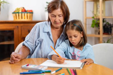 Grandmother watching girl drawing with colored pencil in book at home, showing encouragement and admiration while child focuses on creative activity 