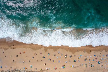 Kumlu deniz kıyısında turistlerin ve renkli şemsiyelerin uzaktan çekimleri, insanların güneşli bir günde kumsalda yaz tatillerinin tadını çıkardıklarını gösteriyor.