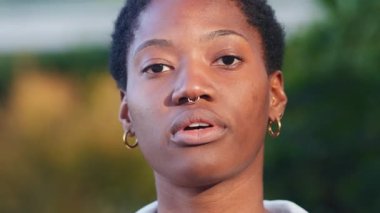 Kısa siyah saçlı, gülümseyen genç siyahi kadının yakın plan portresi açık havada neşeli bir ifadeyle kameraya bakıyor.