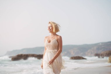 Cheerful blond woman in white summer dress walking near sea at beach enjoying summer vacation with mountains and sky in background during sunny day