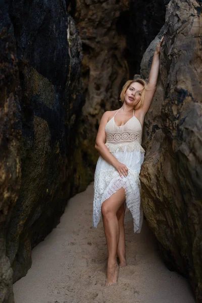 Beyaz dantel elbiseli güzel sarışın kadın kumlu sahilde büyük kayaların arasında poz veriyor. Çekici genç bayan kayaya yaslanırken düşünceli görünüyor.