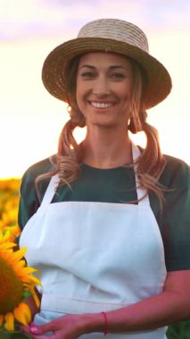 Hasır şapkalı ve önlüklü bir kadın, bir ayçiçeği tarlasında elinde gülümseyerek duruyor. Sıcak günbatımı, açık havanın tadını çıkarırken güzel bir zemin oluşturur..