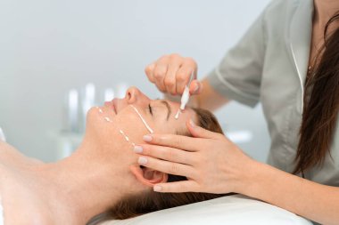Dermatolog, yatakta yatan bir kadının yüzüne krem sürüyor. Genç bayan güzellik salonunda cilt bakımı yaptırıyor. Rahatlatıcı bir tedaviden hoşlanıyor.