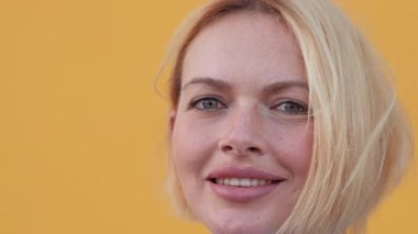 Gülümseyen, kısa sarı saçlı, kendine güvenen ve parlak sarı arka planda pozitif görünen bir kadın portresi. İfadesi profesyonelliği yansıtıyor.