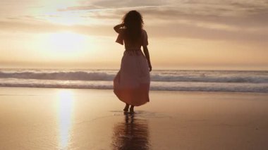 Altın gün batımında kumlu sahilde, okyanus dalgalarına doğru yürüyen ve ılık, huzurlu bir atmosfer yaratan kadın görüntüsü.