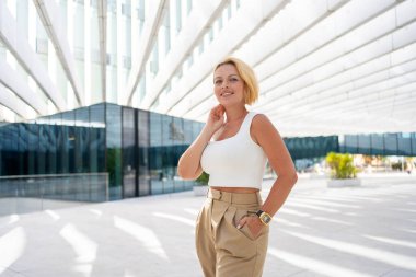 Gündüz vakti profesyonel ve şık görünümlü, modern ofis binasının yakınında gülümseyen, beyaz etekli, kendine güvenen sarışın iş kadını.