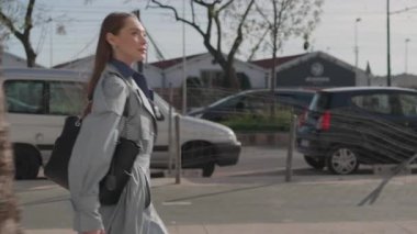 Mavi ceketli genç bir iş kadını şehir kaldırımında kendine güvenen bir ifadeyle yürüyor. Ağaçlar ve yayalar güneşli yaz günlerinde görülebilir.