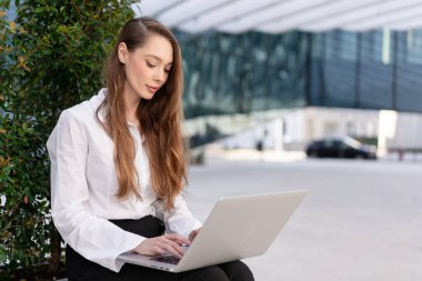 Beyaz gömlekli bir iş kadını modern cam binanın yanında otururken dizüstü bilgisayarda odaklanmış bir ifadeyle çalışıyor.