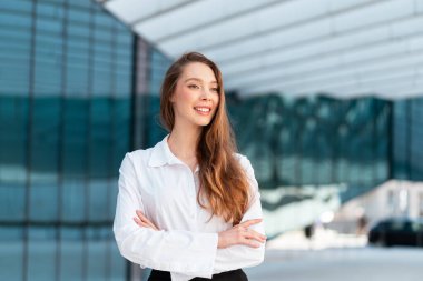 Beyaz gömlekli, kendine güvenen genç bir iş kadını modern cam binanın yanında kollarını kavuşturmuş gülümseyen ve olumlu bir ifadeyle başka tarafa bakıyor.