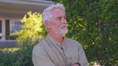 Beyaz saçlı ve sakallı kendinden emin kıdemli işadamı güneşli bir günde yeşillik ve ofis binasının yanında kolları çapraz duruyor.