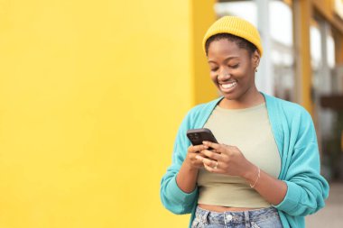 Sarı şapkalı genç bir kadın akıllı telefona bakıyor ve sarı duvara gülümsüyor. Kadın mutlu ve rahat. Pozitif duygu