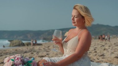 Beyaz yaz elbisesi içinde zarif bir kadın. Kısa sarı saçlı. Kumsalda elinde çiçeklerin yanında şarap kadehiyle oturuyor.