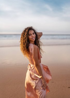 Kıvırcık saçlı, kumlu sahilde gün batımında kameraya bakan gülümseyen genç bir kadın. Özgürlük, neşe ve deniz kenarı mutluluk kavramı