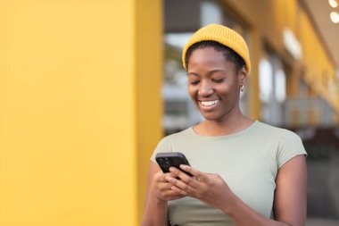 Sarı duvara karşı akıllı telefon kullanan gülümseyen siyah bir kadın. Sarı şapkalı mutlu kadın gündüz vakti telefonla mesaj atıyor.