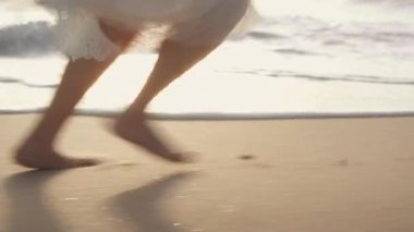 Kumlu sahilde çıplak ayakla koşan beyaz elbiseli bir kadının güneş batarken okyanus dalgalarının yanında ayak izleri bırakması.