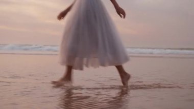 Güneş batarken kumlu sahilde çıplak ayakla koşan beyaz elbiseli genç bir kadın neşeli ve tasasız bir atmosfer yaratıyor.