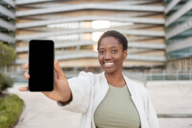 Gülümseyen siyah kadın, bulanık binaya karşı boş ekranla akıllı telefon gösteriyor. İlan, uygulama veya web sitesi için cep telefonu görüntüleme şablonu