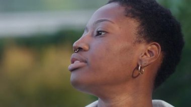 Siyahi bir kadının dışarıda sigara içerkenki yakın çekimi. Genç bir kadın, arka planda bulanık bir şekilde kameraya bakarken duman üflüyor.