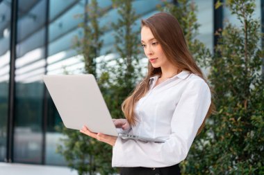 Modern ofis binasının yanında duran beyaz gömlekli iş kadını dizüstü bilgisayar kullanıyor. Profesyonel teknoloji ve kurumsal iş kavramı