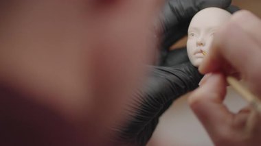 Eldiven giyerken ince fırça kullanarak bebek kafası çizen ressamın yakın çekimi, bebek oluşturma sürecindeki hassas ayrıntıları gösteriyor