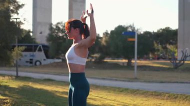 Dışarıda egzersiz yaparken direnç bandı geren bir kadın. Kadın sporcu, şehirde köprü yakınlarında gün ışığında çimlerde egzersiz yaparken kollarını hareket ettiriyor.