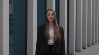 Modern binanın önünde duran siyah ceketli, gülümseyen genç bir kadının görüntüsünü yakınlaştırın, şehir ortamında kendine güvenen bir ifadeyle kameraya bakın.