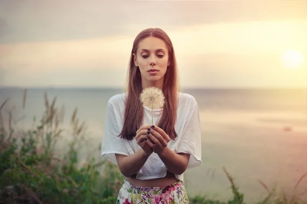 Portrait of a brunette young woman  with a big dandelion on a background of warm sunset. summer, outdoors. — Stock Photo, Image