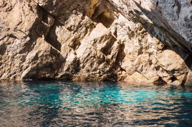 Capri, İtalya - 04.29.2025: Capri adasının kıyısındaki kayalıkların manzarası; kıyıya yakın açık mavi deniz suyu güzel aydınlatılır..