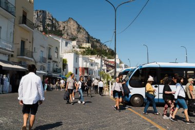 Capri, İtalya - 04.29.2025: liman yakınlarındaki otobüslerde halk ve turistlerin bulunduğu bir şehir caddesinde günlük durum; evler, dağlar ve mavi gökyüzü görülebilir.