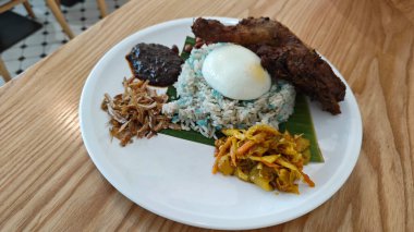 a plate of Blue Butterfly Pea nasi lemak rice with fried chicken drumstick,pickle vegetable,anchovies,peanuts and sambal. 