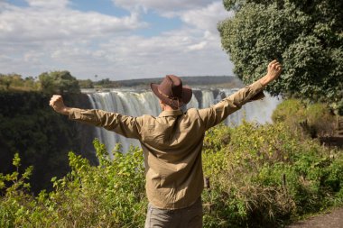 a man in a cowboy hat stands on a cliff against the background of Victoria Falls with his arms outstretched. rear view clipart