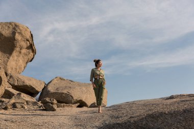 Khakki renkli yürüyüş kıyafetleri giyen bir kız mavi gökyüzüne karşı bir kayanın üzerinde duruyor..