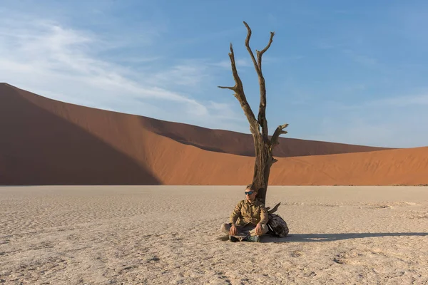 A hiker in hiking clothes and a backpack sits in the middle of Dead Vlei by a withered tree against the backdrop of a dune and blue sky. Sossuflei, Namibia