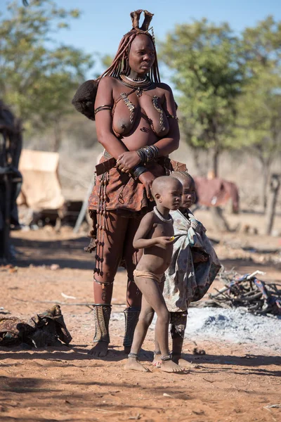 Namibya, Afrika, 18 Temmuz 2019. Himba 'lı bir kadın, geleneksel üstsüz elbisesiyle çocuklarının yanında duruyor..