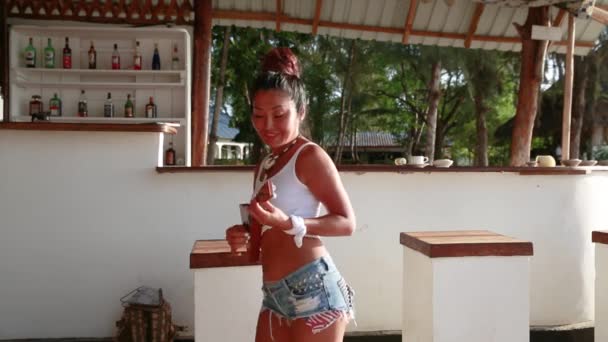 Сексуальна дівчина азіатського походження в коротких шортах з денімом, а балаїка в руках танцює на пляжі в барі. — стокове відео