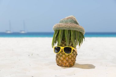 Olgun ve sulu bıyıklı ananas, hippi şapkası ve plajda güneş gözlüğü, arka planda gök mavisi deniz.