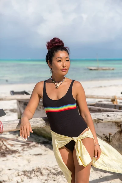 Ein schönes Mädchen in einem schwarzen Badeanzug und einem Verband an der Hüfte steht neben einem alten Fischerboot am Strand — Stockfoto