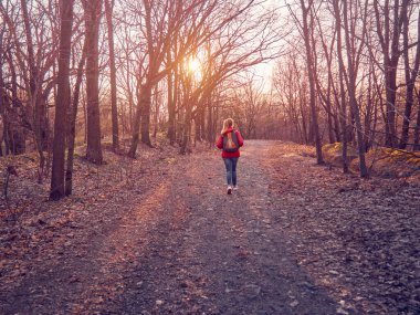 Sırt çantalı bir kız batan güneşin altında yapraklı bir sonbahar parkında yürüyor.
