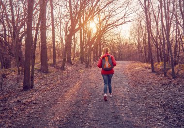 Sırt çantalı bir kız batan güneşin altında yapraklı bir sonbahar parkında yürüyor.