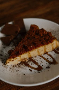 Lezzetli bir dilim kahveli cheesecake kakaoyla kaplı beyaz tabakta çikolata ve koyu ahşap arka planda karamel.