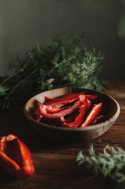 Ahşap tabakta parlak kırmızı biberler ve mutfaktaki koyu masa arkasında yeşil dereotu. Lezzetli olgun sebzeler yakın plan pişirmeye hazır. Hungurian leçosu için girdaplar.